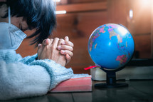 person wearing a face mask with praying hands beside of a globe 