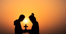 silhouettes of two people holding a cross and praying at sunset 