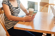 a woman sitting at a table texting and holding a coffee cup 