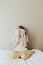 A young woman sitting on a bed and reading a book.