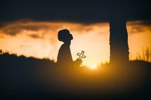 Young man praying and holding Cross under a tree at sunset background.