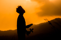 silhouette of a boy holding a cross and Bible outdoors 