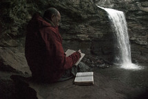 man writing in a journal and reading a Bible near a waterfall