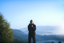 a man in a coat standing on a mountaintop praying 