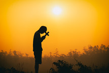 silhouette of a man praying at sunset holding a cross 