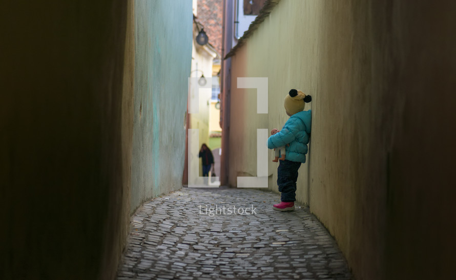 toddler girl holding a doll standing in an alley 