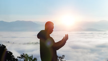 a man in a coat standing on a mountaintop in the clouds praying 