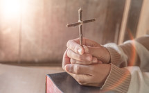 praying hands holding a cross over a Bible 
