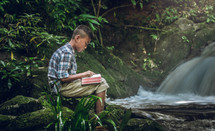 a boy reading a Bible outdoors near a waterfall 