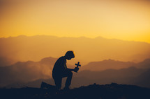 a silhouette of a boy holding a cross and kneeling in prayer at sunset 