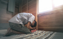 man kneeling on a bed by a window praying 