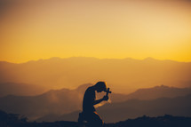 silhouette of a boy kneeling in prayer on a mountaintop at sunset 