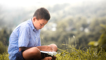 a boy sitting outdoors reading a Bible 