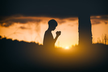 a silhouette of a boy praying outdoors at sunset 