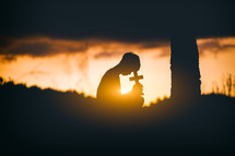 Young man praying and holding Cross at sunset 