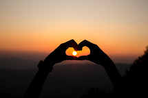 hands forming a heart around a sun at sunset 