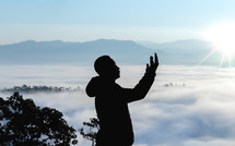 a man standing on a mountaintop in the clouds with raised hands 