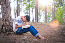 a boy sitting under a tree reading a Bible and praying 