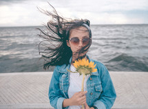 a young woman standing at the shore holding a sunflower 