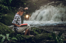 a boy reading a Bible outdoors near a waterfall 