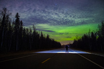 man with a flashlight standing in the middle of a road under an aurora 