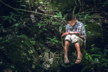a boy sitting in a forest on a mossy rock reading a Bible 
