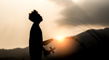 silhouette of a boy holding a cross and Bible outdoors 
