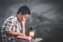 a boy reading a Bible outdoors by a lantern 