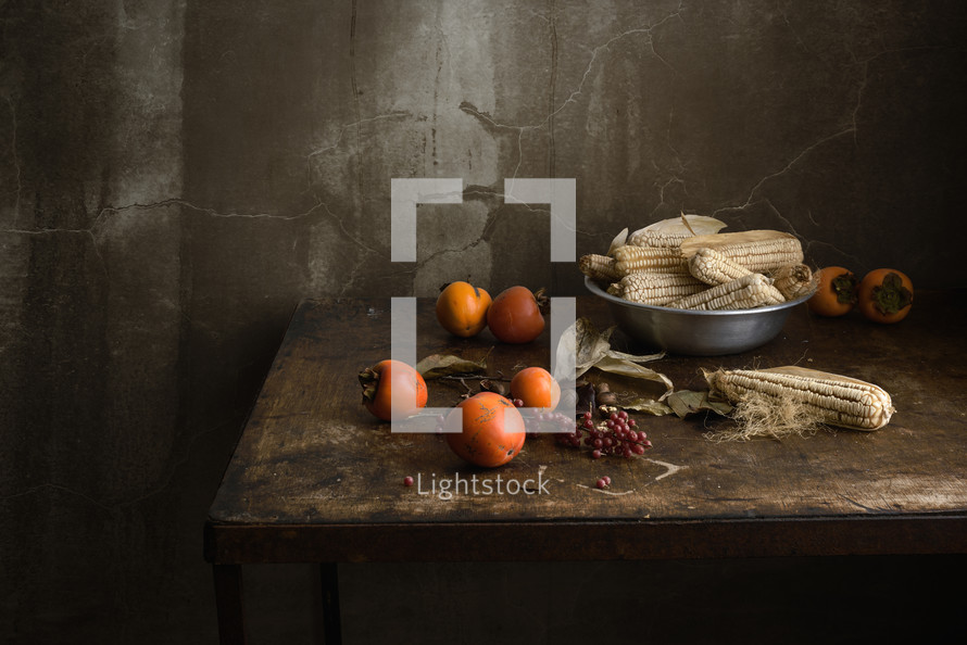 A rustic setting of a table with fruit and ears of corn.