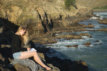 a woman sitting on rocks along a shore writing in a journal 