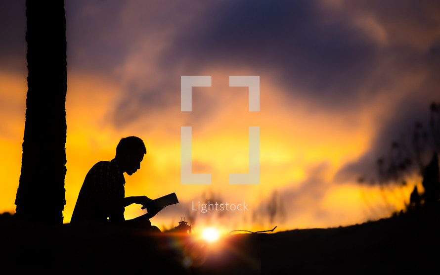 silhouette of a boy reading a Bible outdoors at sunset 