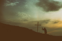 man kneeling in prayer next to a cross with a dramatic sky 