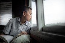 a boy reading a Bible and looking out a window 