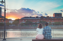 a couple sitting on a bench looking out at a river at sunset 