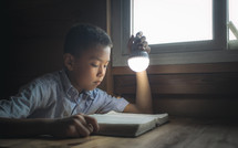 a boy reading a Bible by a window holding a light 