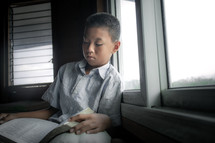 a boy reading a Bible in a window 