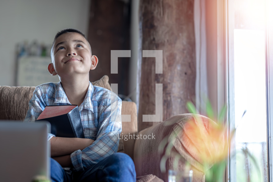 a boy holding a Bible and sitting in front of a computer at home 