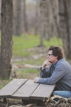 A man sits at a table in the woods.