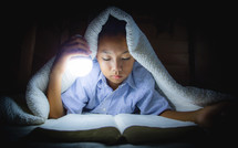 a boy reading a Bible under a blanket 
