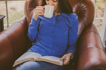A young woman sitting in a chair reading the Bible and drinking coffee