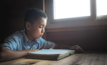 a boy sitting by a window reading a Bible 