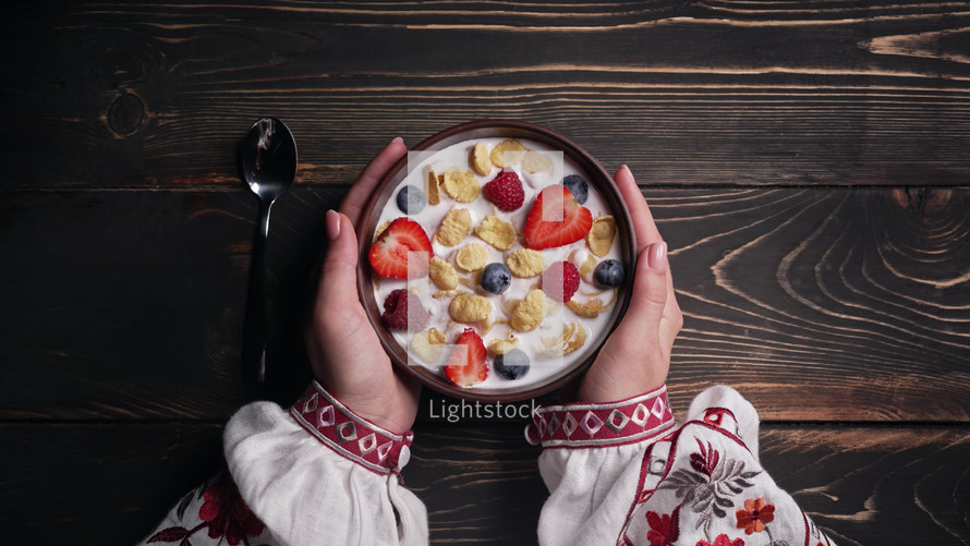Female hands with breakfast - cereal with milk and berries. Ukrainian clay bowl on wooden table. Woman in traditional embroidered shirt. Top view. Healthy food, eating, great morning