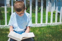 a young boy sitting in grass on a sunny day reading a Bible in solitude with his head bowed 