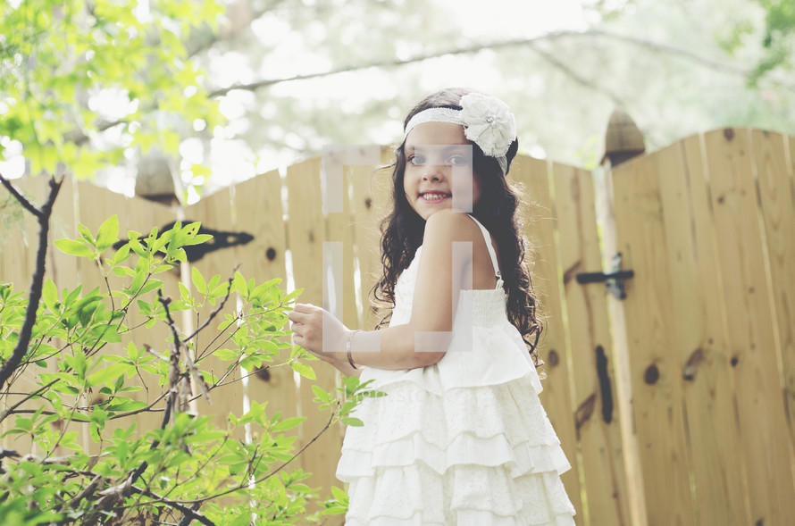 A little girl in a white dress standing by a wooden fence.