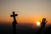 raised hands holding up a cross with sunlight at sunset in the background