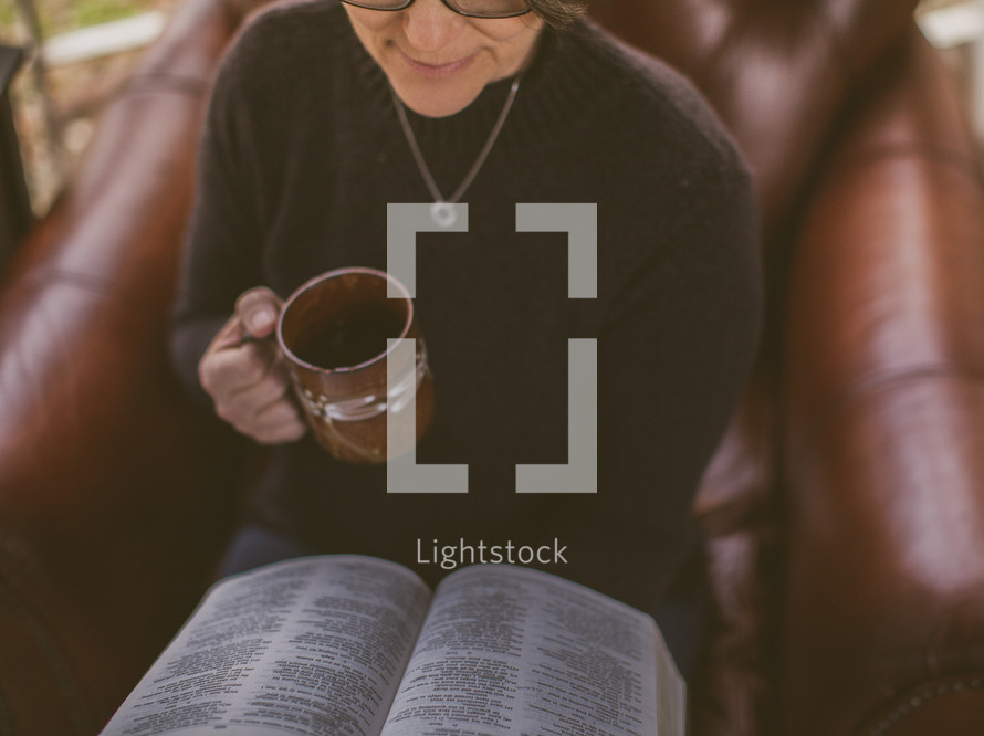 Woman sitting in a chair reading the Bible and drinking coffee.