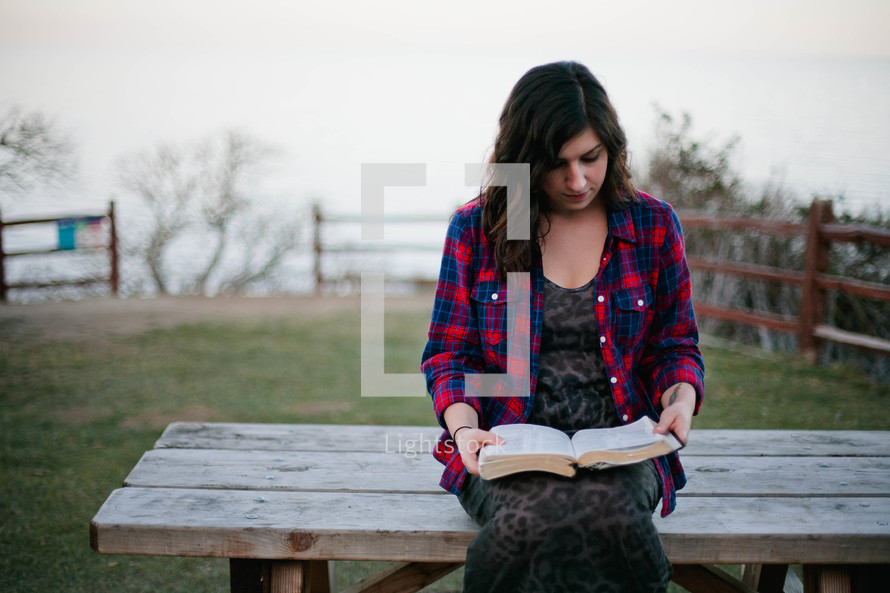 A woman sitting on a bench outdoors reading a Bible. 
