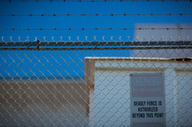barbed wire and a chain link fence, forbidden area, deadly force authorized beyond this point, no trespassing, warning, sign