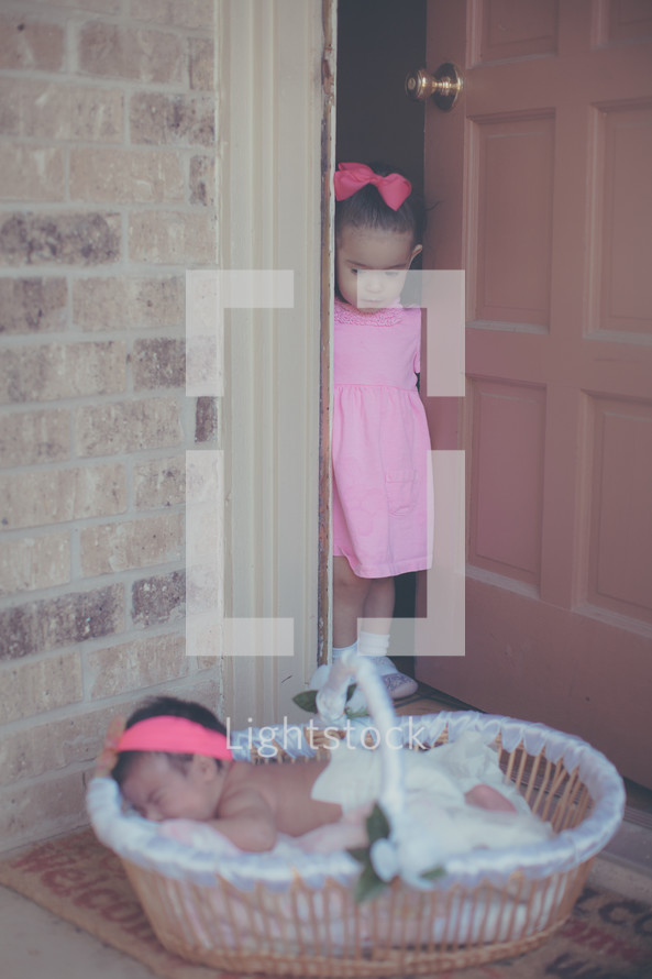 a young door looking out a door at her newborn sister in a basket 