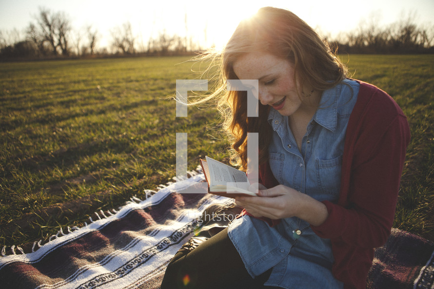 young woman sitting on a blanket in the grass reading a pocket Bible under sunlight